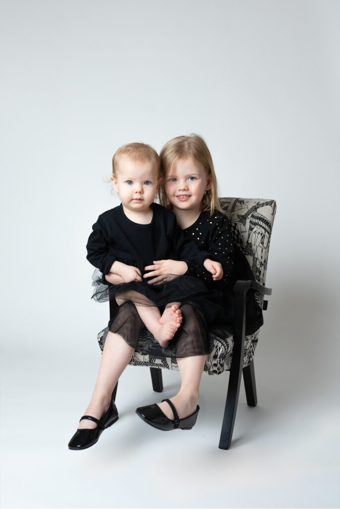 Lillasyster sitter i knät på storasyster i en fin fåtölj i en fotostudio